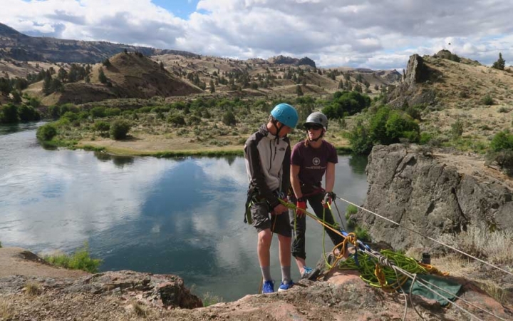 Two people wearing safety gear are secured by ropes near the edge of a cliff. One person appears to be an instructor, giving direction to the other person. There is a lake below them.